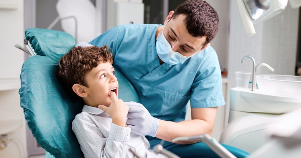 A képen egy gyerek ül a fogorvosi székben, a szájába mutat, a fogorvos pedig belenéz a szájába