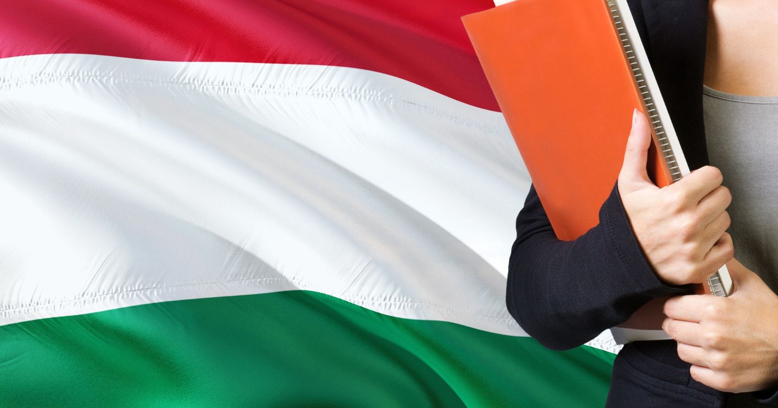 A képen a magyar zászló előtt áll egy ember, kezében mappával