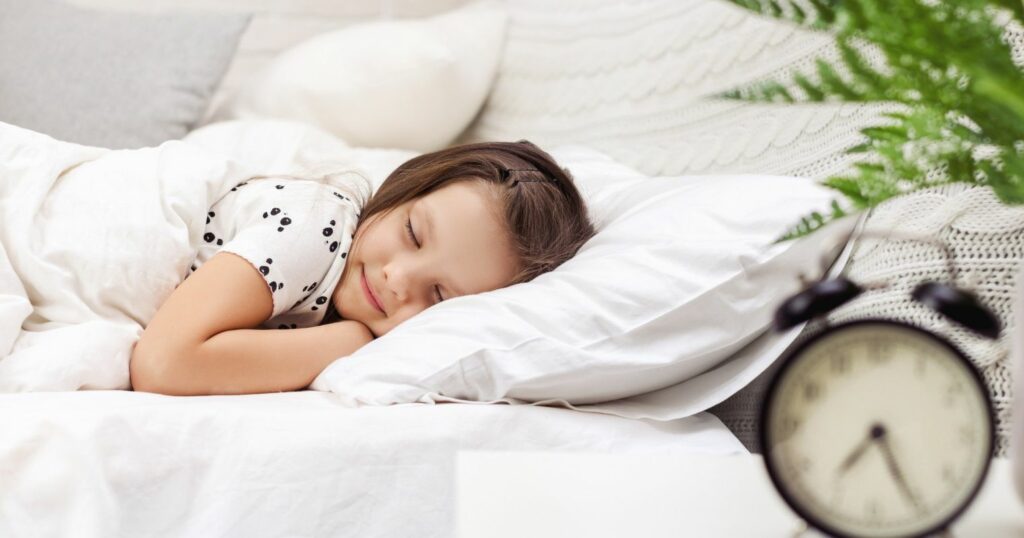 A képen egy kislány az ágyában alszik, a kép előterében egy vekker látható