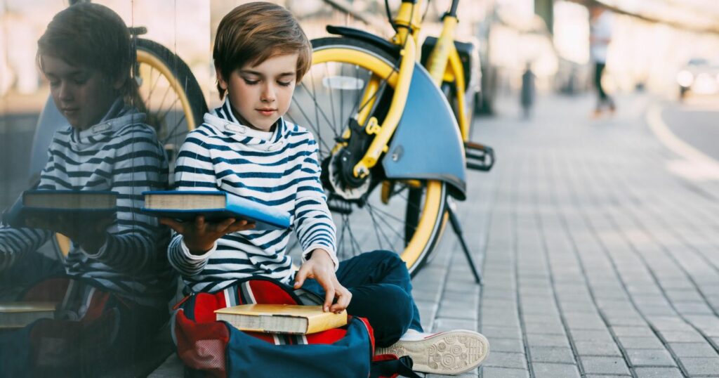 A képen egy kisfiú ül a földön, és az iskolatáskájába könyveket pakol