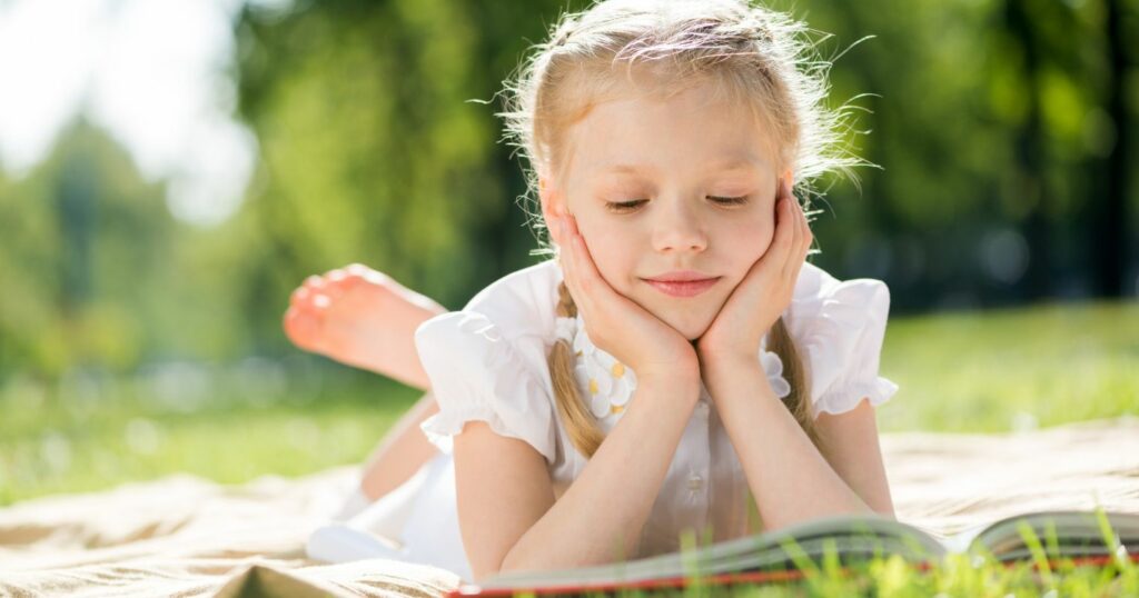 A képen egy kislány könyvet olvas a fűben