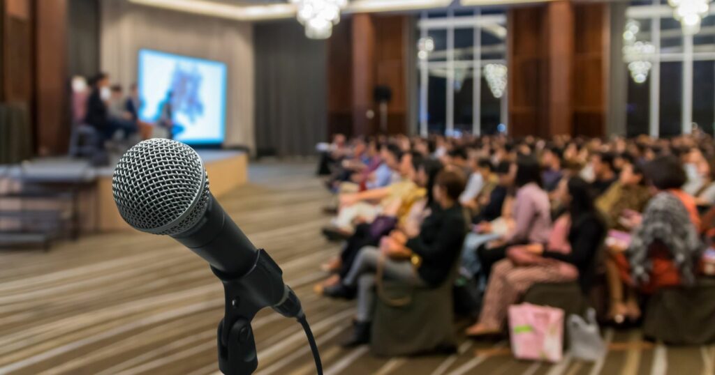 A képen egy mikrofon látszik, a háttérben egy konferencia látható