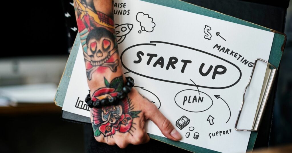 A képen egy tetovált kar tart egy mappát, rajta a startup felirat olvasható