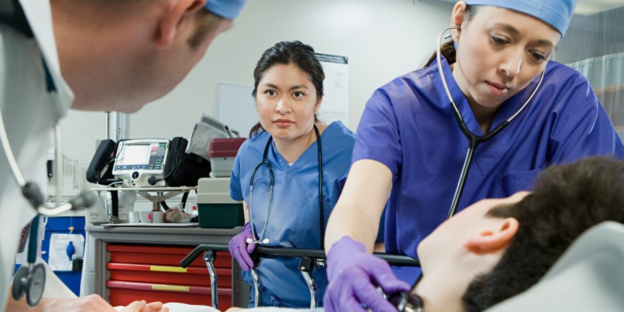 Három egészségügyi dolgozó áll körül egy fekvő beteget, az egyik a nyakán tartja a szetoszkópját.