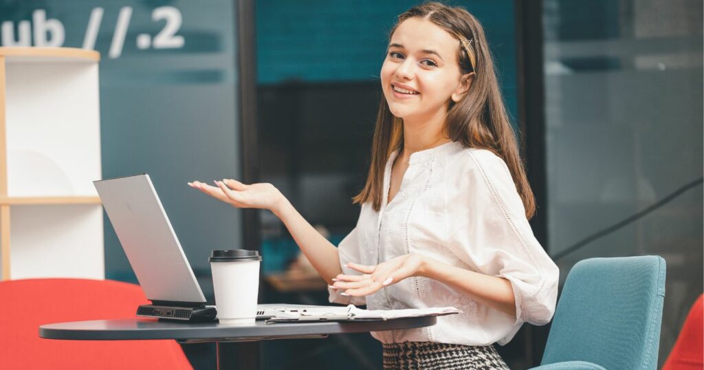 A képen egy mosolygó fiatal lány látható asztalnál ülve, laptoppal.