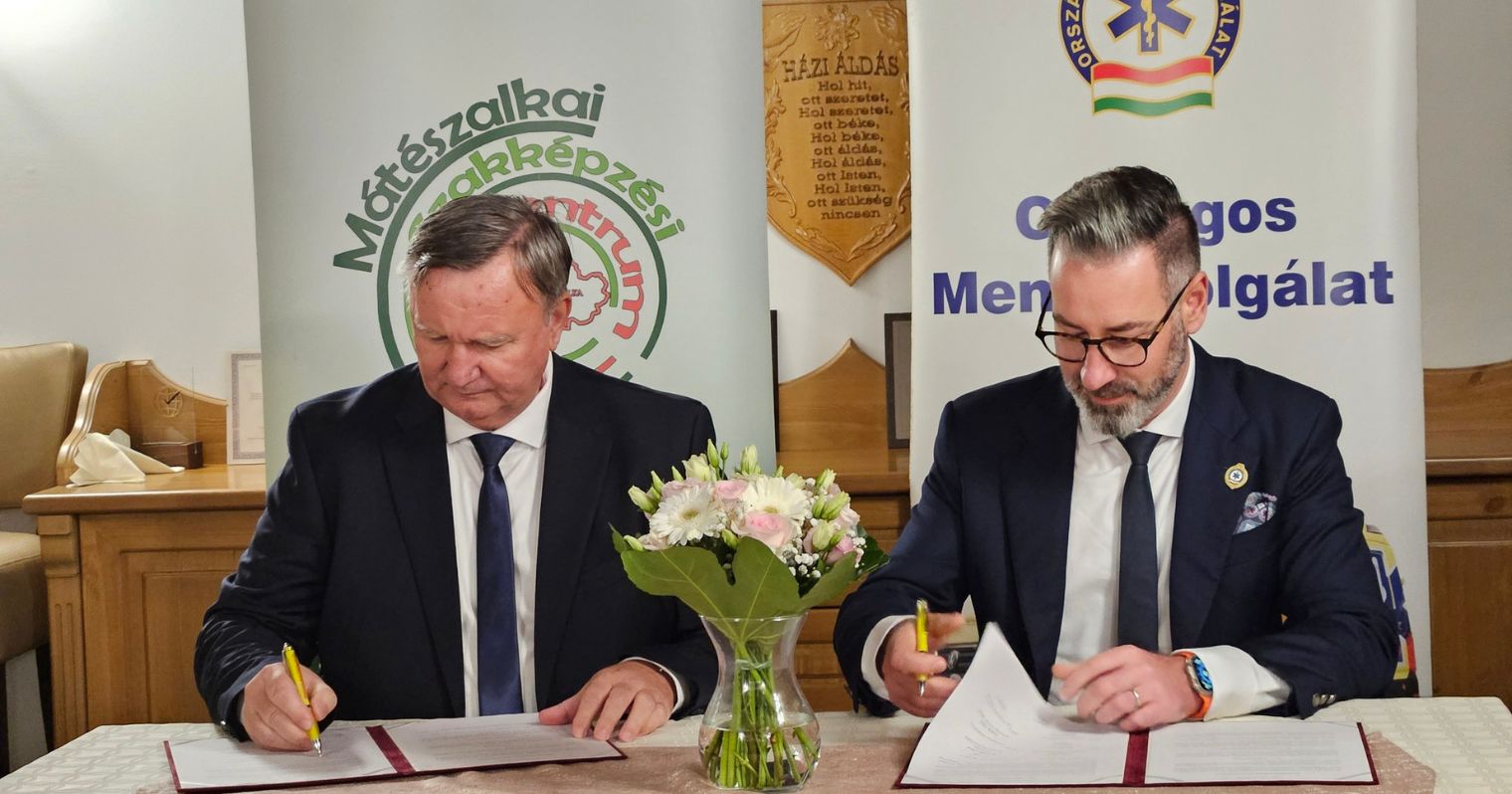 Csató Gábor, az OMSZ főigazgatója és Rostás János Tibor, a Mátészalkai Szakképzési Centrum kancellárja aláírják az együttműködést a két intézmény bannerei előtt egy asztalnál.