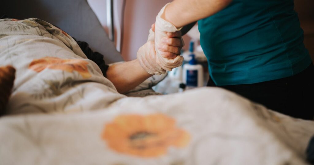 A képen egy ágyban fekvő ember fogja meg az ápolója kezét