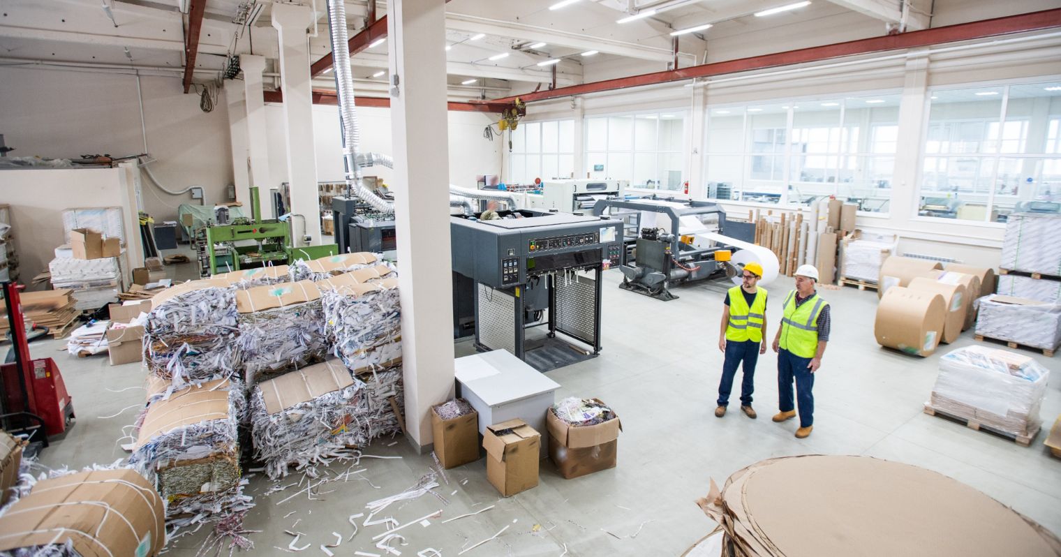 Papír újrahasznosító gyár dolgozói védőmellényben állnak csomagolt papírok és, vágógépek mellett.