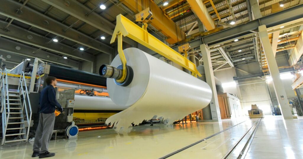 Nagy papír henger egy gyárban, aminek a mozgatását egy munkás irányítja.