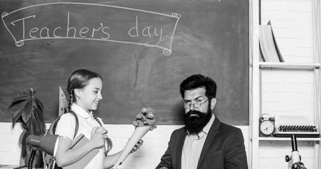 A képen egy kislány ad virágot egy tanárnak, a háttérben egy táblára a pedagógusnap szó van felírva angolul