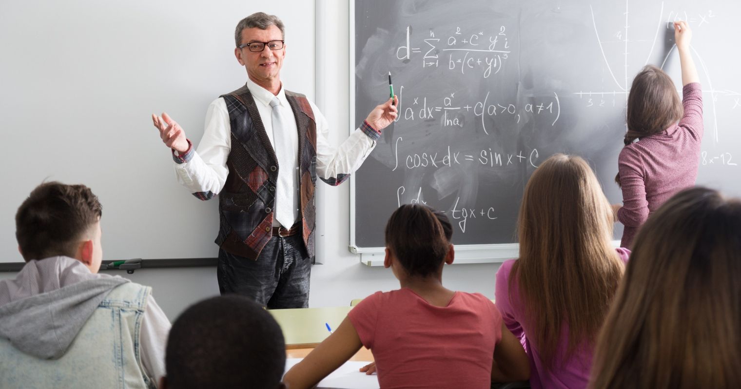 A képen egy tanár áll a tábla előtt, körülötte osztályteremben tanulók ülnek
