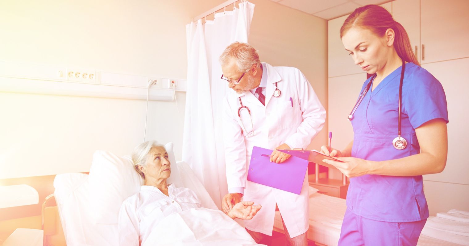 Idős beteg ágya mellett áll az orvos, mellette pedig egy nővér mappával a kezében jegyzetel.