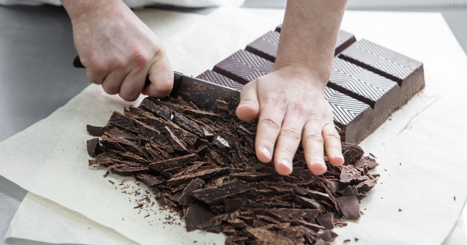 Egy óriási tábla csokit vág kis darabokra egy nagy késsel egy férfi, a képen csak a kezei látszanak.