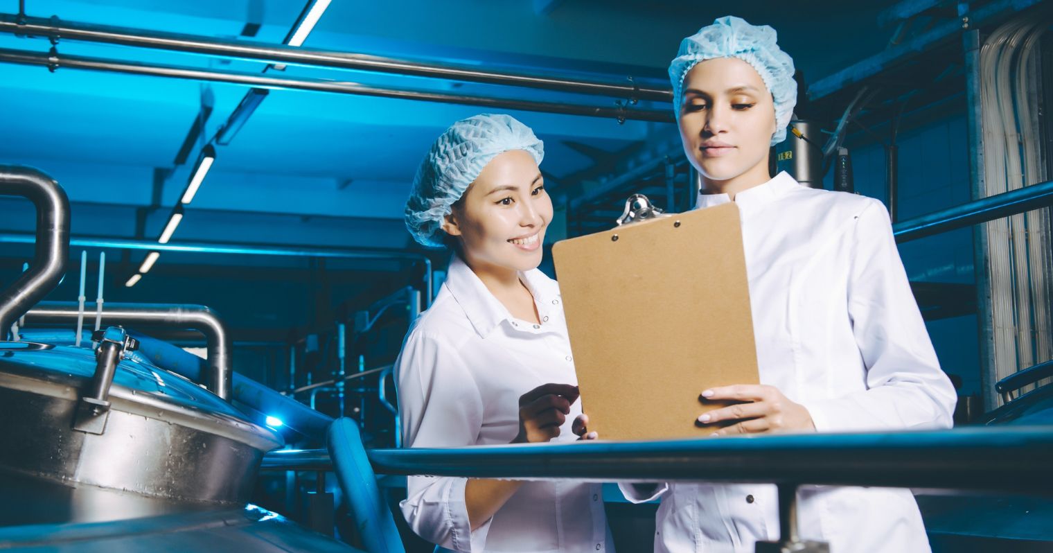 Kék világítású gyárban két hajhálós és fehér köpenyes nő egy írótáblát néznek.
