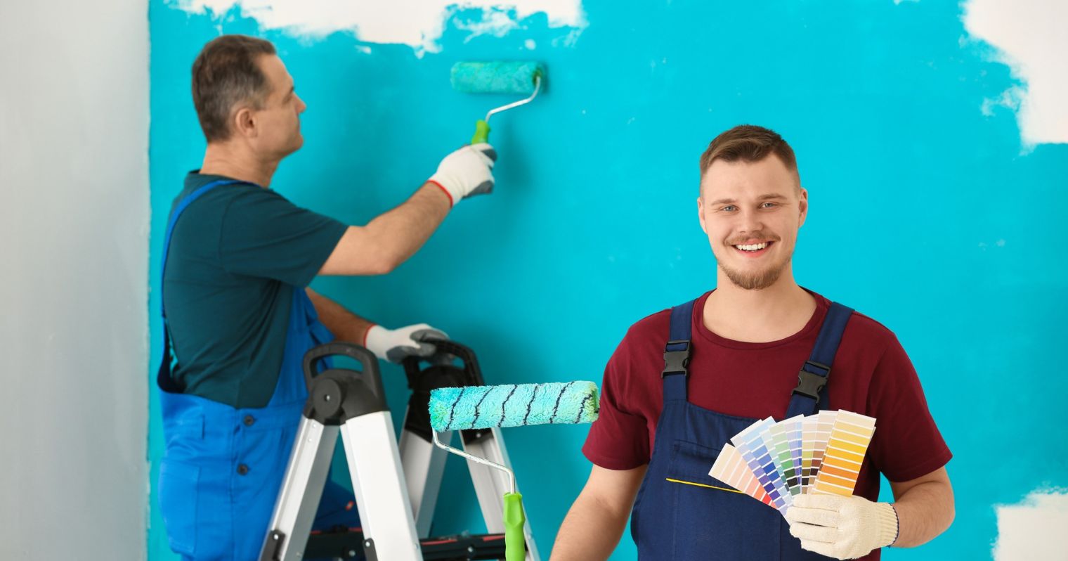 Két festő épp kékre fest egy falat, az egyik egy létrán állva fest, a másik színmintákat és festőhengert a kezében fogva a kamerába mosolyog.