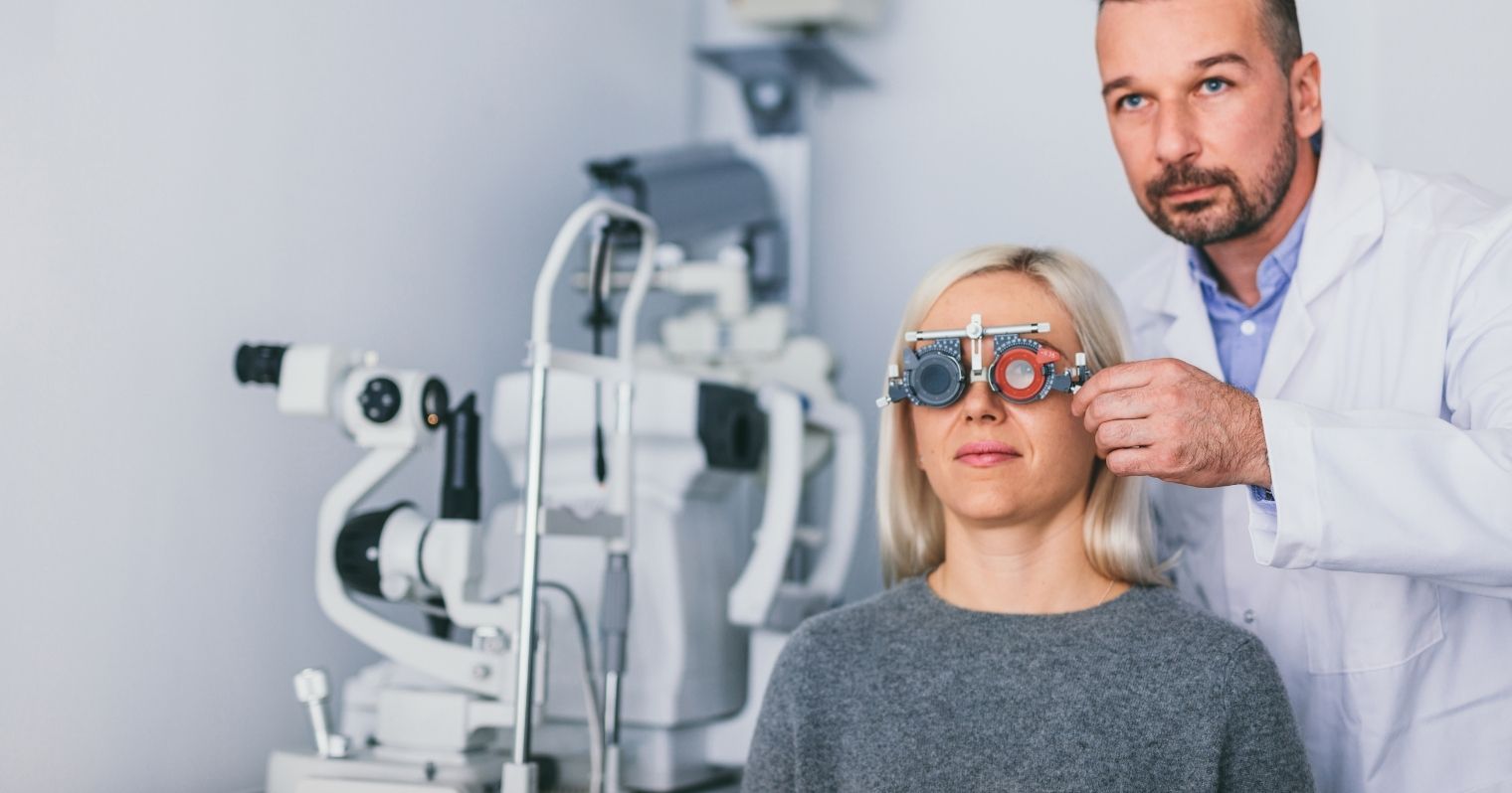 Orvosi köpenyes optikus férfi lencséket tart egy nő szeme elé, a háttérben egy bonyolult orvosi műszer látható.