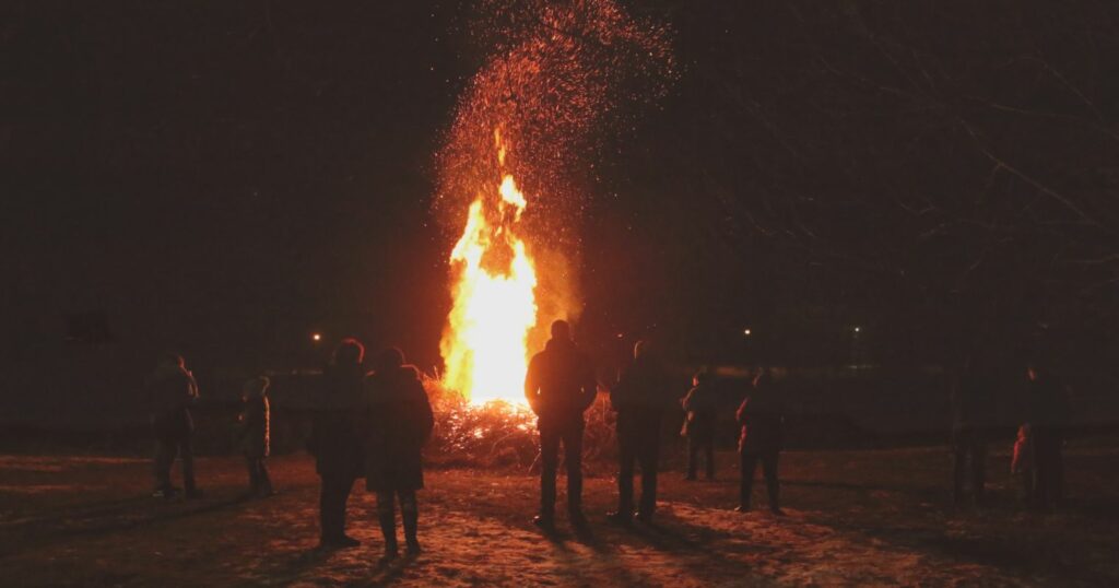 Téli környezetben, sötétben, kabátos emberek egy nagy tüzet állnak körbe.
