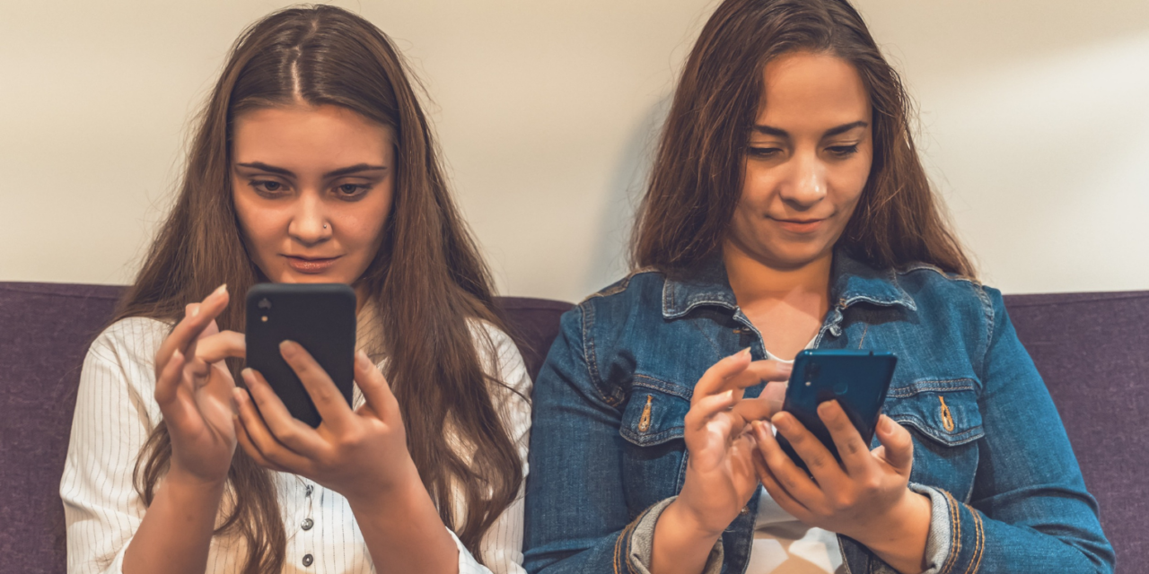 Két fiatal lány egymás mellett ül a kanapén, mindketten a telefonjukat nyomkodják.