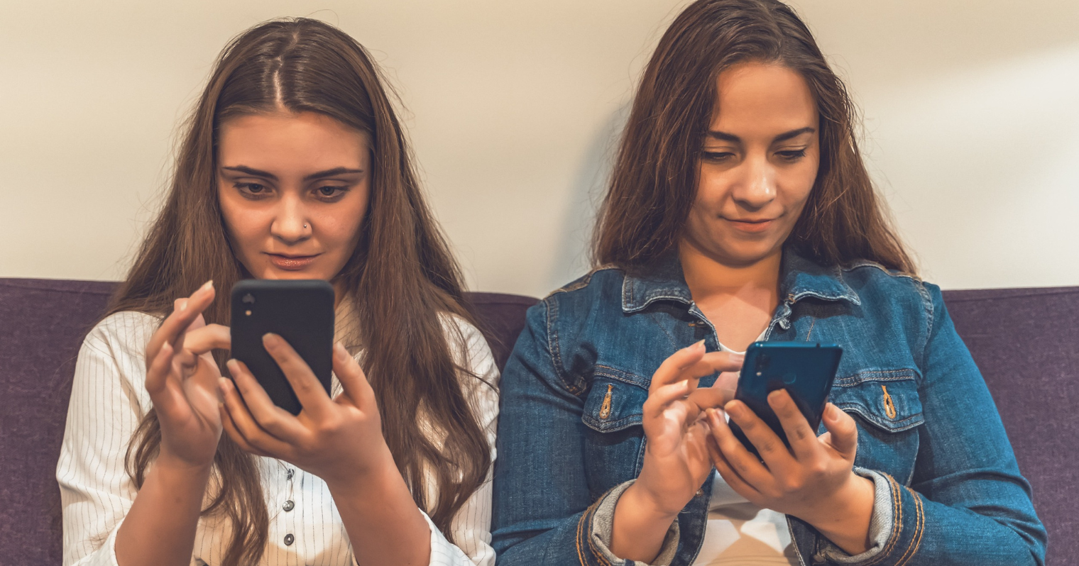 Két fiatal lány egymás mellett ül a kanapén, mindketten a telefonjukat nyomkodják.