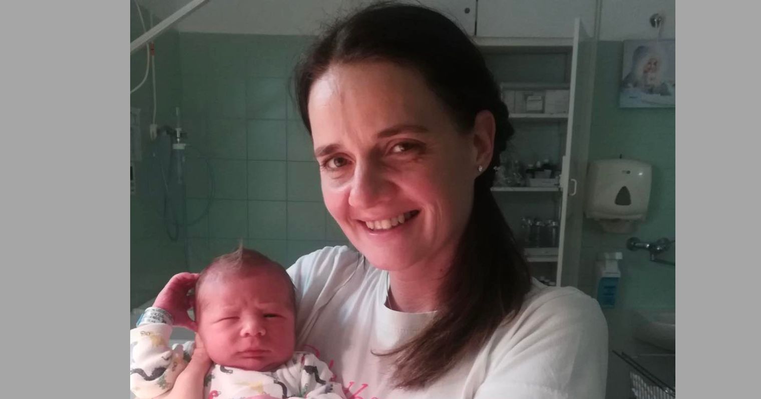 Csanádiné Bartha Ágnes csecsemő- és gyermekápoló áll egy kórházi szobában kezében egy újszülött babával.