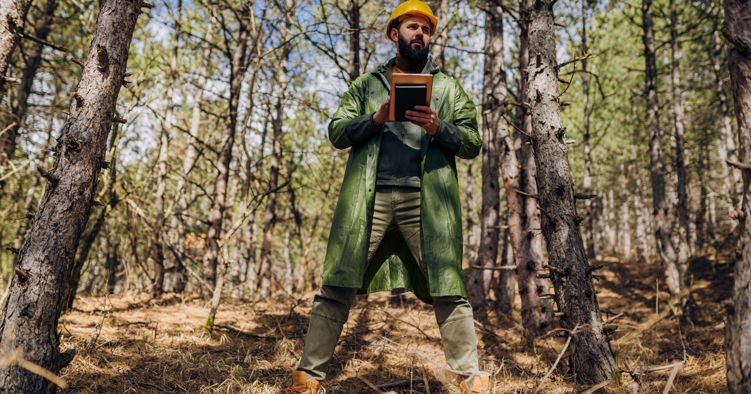 Zöld ruhában áll az erdőben egy ember, kezében jegyzetfüzettel.