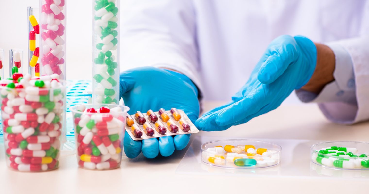 Kék gumikesztyűs kezeken tart színes gyógyszereket egy ember, mellette az asztal sok másik színes gyógyszer van üvegekben.