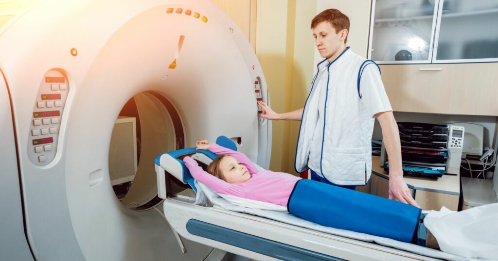 CT gépben egy kislány fekszik és egy egészségügyi asszisztens épp elindítja a gépet.