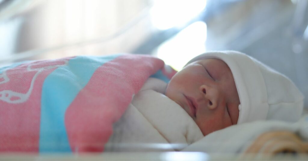 Újszülött csecsemő takaróban és sapkában a kórházi ágyában alszik. Csecsemő- és gyermekápoló szakmához kapcsolódó kép.
