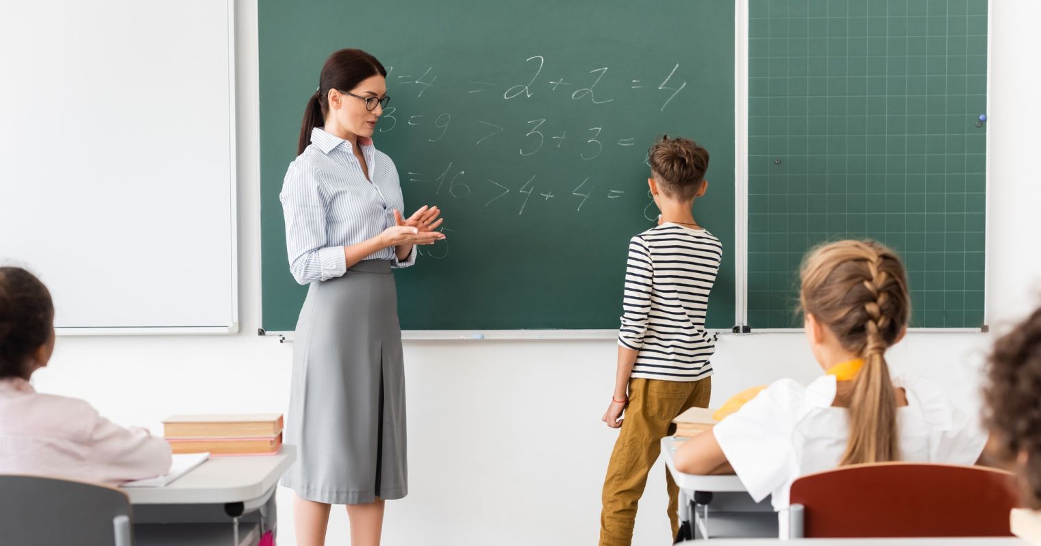 A képen egy pedagógus és gyerekek láthatók a tábla előtt, tanítás közben.