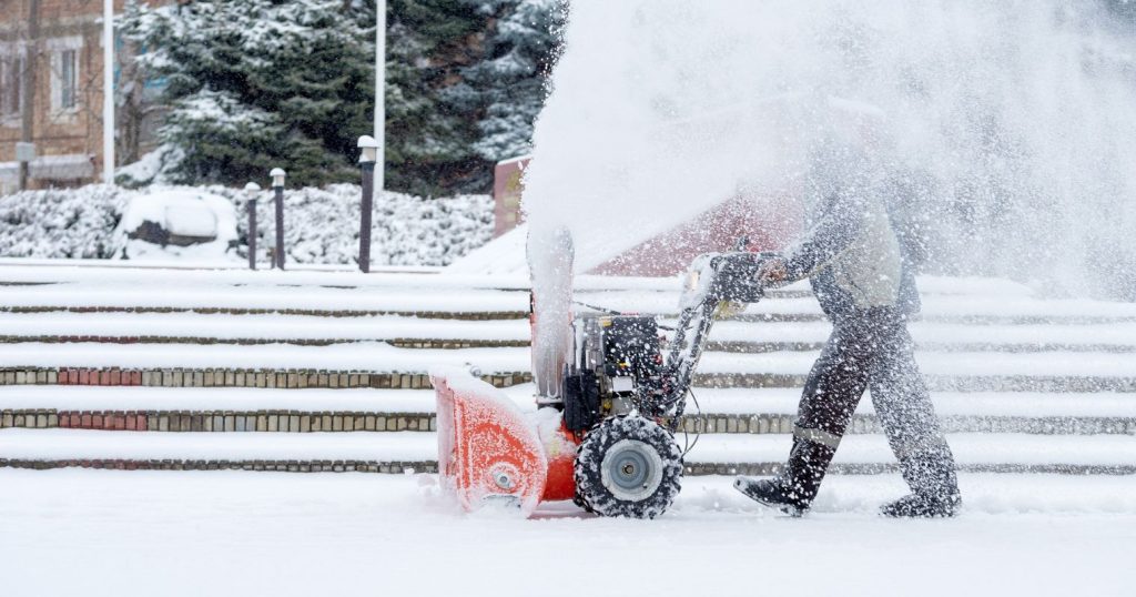 Havas területen hóeltakarító gépet tol egy ember, közben pedig a hó az arcába fújódik.