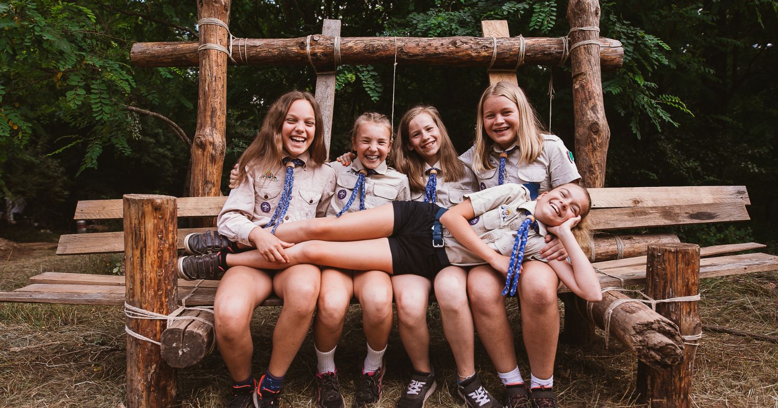 Cserkész egyenruhás lányok ülnek egy kötözött fa szerkezeten csoportképhez.